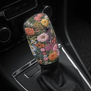 May include: A floral patterned gear shift knob with a black leather base. The pattern features pink, red, yellow, and white flowers on a dark green background.