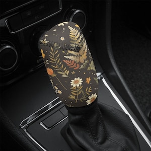 May include: A shift knob cover featuring a dark brown backdrop adorned with a floral design. The pattern showcases white daisies, orange blooms, and an assortment of green and brown leaves. The cover is fitted on a black gear stick.
