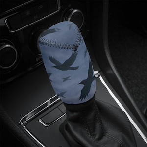 Könnte beinhalten: Ein blauer Schalthebelbezug mit einem Muster aus fliegenden Krähen. Der Bezug ist zylindrisch und passt über den Schalthebel, mit einer schwarzen Naht oben. Der Hintergrund ist ein gedämpftes Blau.