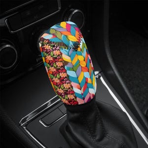 May include: A patterned car gear stick cover. The cover is decorated with a floral and braided leaf design in a variety of colours, including red, orange, yellow, blue, and green, adding a decorative touch to the car's interior.