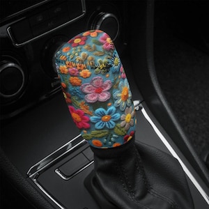 May include: A colourful floral patterned gear shift knob cover. The cover is made of a soft, textured material and features a variety of flowers in shades of pink, blue, yellow, and green. The cover is designed to fit over a standard gear shift knob.