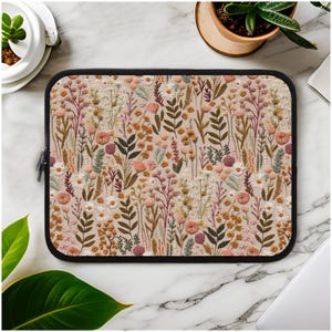 Puede incluir: Una funda para portátil con un patrón floral. El patrón presenta una variedad de flores y hojas en tonos de rosa, verde y marrón. La funda está hecha de un material suave y duradero y tiene un cierre de cremallera.