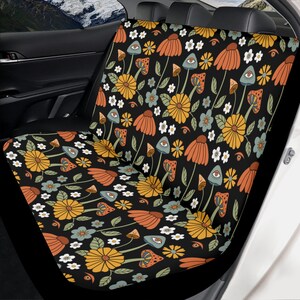 May include: A black car seat cover featuring a repeating pattern of orange and yellow flowers, green leaves, small white blossoms, and mushrooms with eyes. The fabric covers the backrest and seat cushion of a vehicle's rear bench seat.