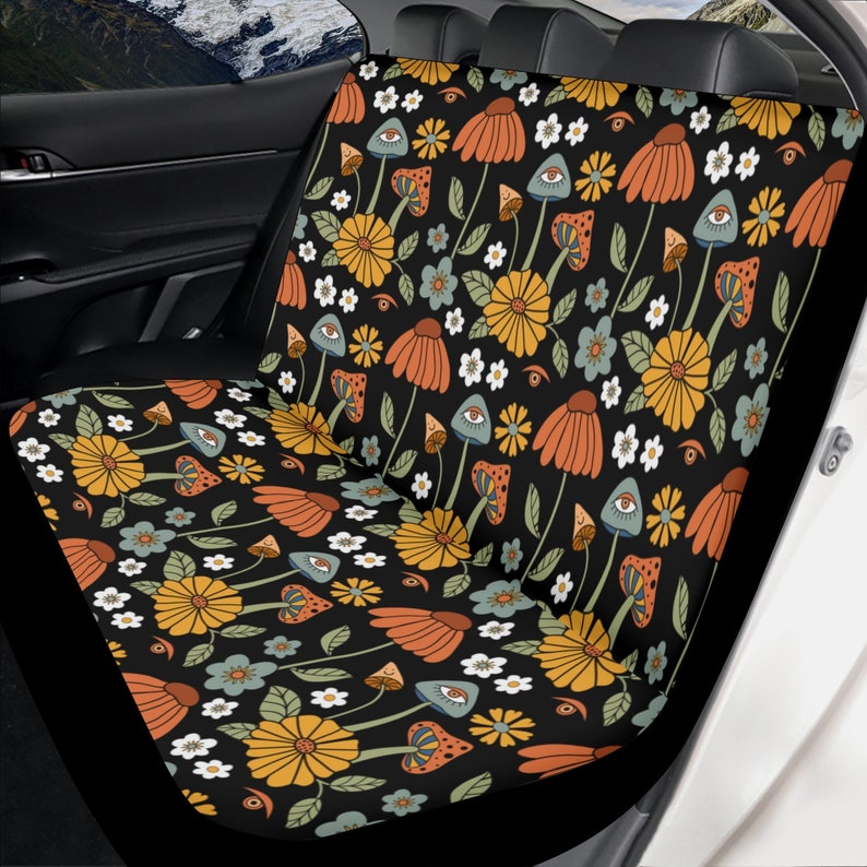 May include: A black car seat cover featuring a repeating pattern of orange and yellow flowers, green leaves, small white blossoms, and mushrooms with eyes. The fabric covers the backrest and seat cushion of a vehicle's rear bench seat.