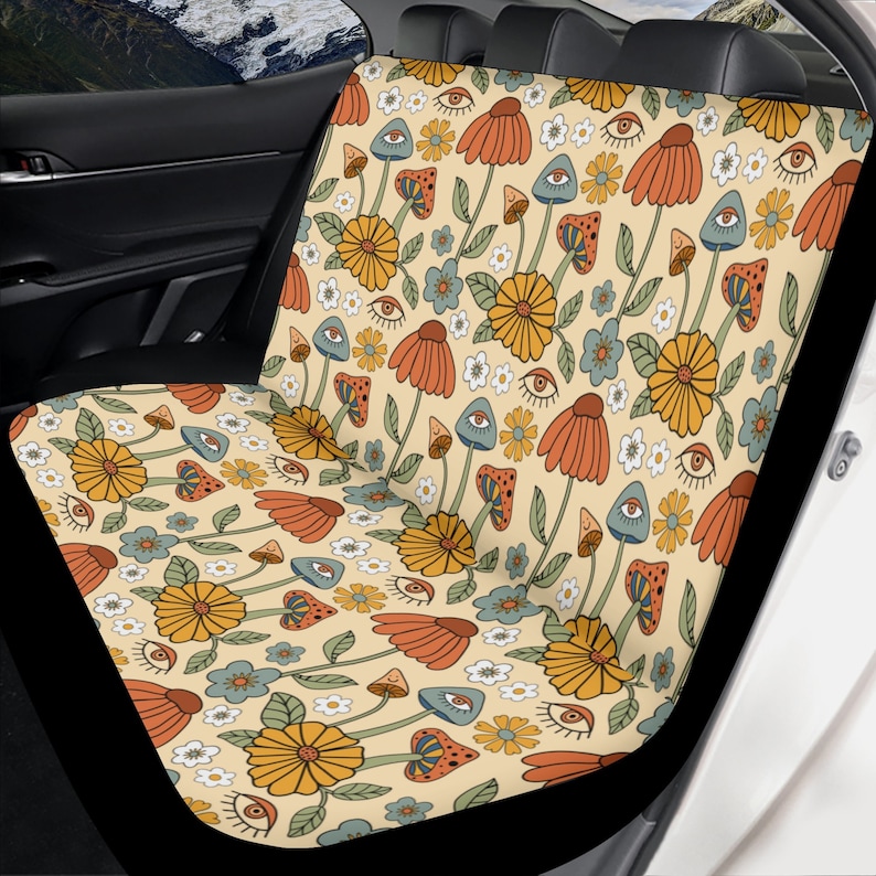 May include: A car seat cover featuring a repeating pattern on a cream background. The design includes orange and yellow flowers, blue and orange mushrooms, small white flowers, and stylized eyes with eyelashes in a consistent graphic print.