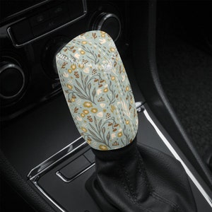 May include: A floral pattern shift knob cover in a car interior. The cover features a light blue background with a repeating pattern of brown, tan, and white flowers and leaves. The cover is made of a soft, stretchy material.