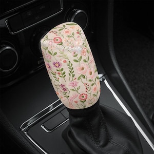 May include: A floral patterned car gear shift knob with a beige background and pink and white flowers. The knob is attached to a black leather gear shift boot.