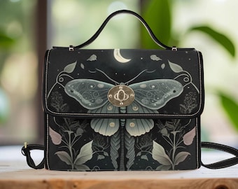 May include: Black satchel bag featuring a large moth design with a crescent moon and floral accents. The bag has a gold-colored clasp, a top handle, and a detachable shoulder strap. The moth is in shades of gray and teal.