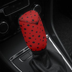 May include: A red gear stick cover featuring a pattern of black polka dots. The cover is made from a soft fabric and is designed to fit over the gear stick in a car. A decorative and stylish car accessory.