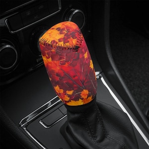 May include: A car gear stick cover with a design of autumn leaves. The cover displays a pattern of red, orange, and yellow leaves, with black stitching at the top. The cover is designed to fit over the gear stick lever.