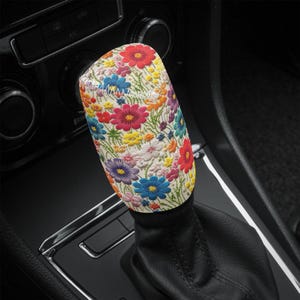 Op de afbeelding: Een witte versnellingspookknop met een kleurrijk bloemenpatroon. Het patroon heeft roze, rode, gele, blauwe en groene bloemen op een witte achtergrond.
