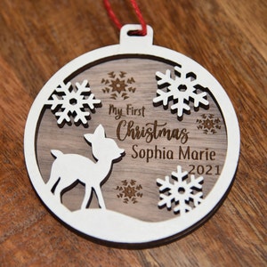 May include: A white wooden Christmas bauble featuring a deer silhouette, snowflakes, and the text "My First Christmas Sophia Marie 2021".