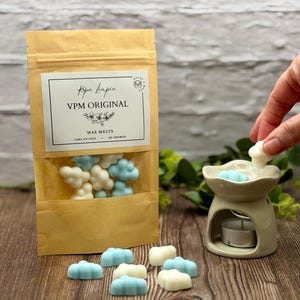 Puede incluir: Una bolsa de papel kraft con ventana transparente muestra ceras aromáticas en forma de nube en blanco y azul claro. La bolsa lleva la etiqueta "VPM ORIGINAL" con "Wax Melts" y "Cera de Soja". Un quemador de aceite de cerámica contiene una cera, con más sobre una superficie de madera.
