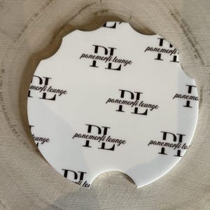 May include: White car coaster with a unique scalloped edge. The coaster features a repeating pattern of the text "panemorfi lounge" in black. The coaster has a cutout for easy removal from a car cup holder.