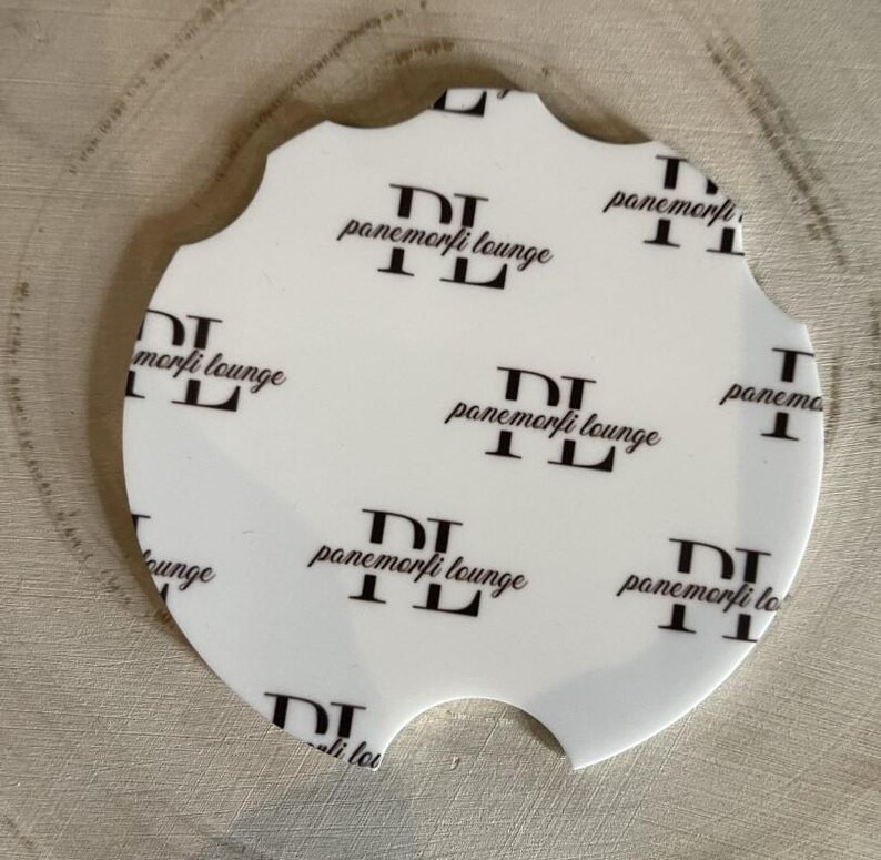 May include: White car coaster with a unique scalloped edge. The coaster features a repeating pattern of the text "panemorfi lounge" in black. The coaster has a cutout for easy removal from a car cup holder.