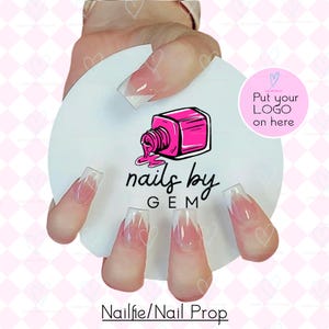May include: A white nail prop with the text "nails by GEM" and a pink nail polish bottle graphic. The prop is holding several artificial nails with a pink and white design. The text "Nailfie/Nail Prop" is at the bottom.