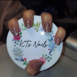 May include: Close-up of a hand with glittery rose gold nail polish. The nails are painted with a sparkling finish. The hand is holding a white circular object with the text "Kts Nails" and a floral design.