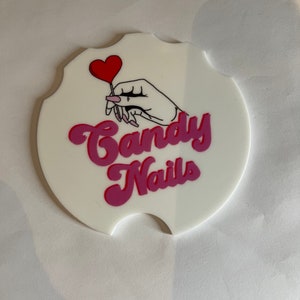 May include: White car coaster with a scalloped edge, featuring a hand holding a red heart-shaped lollipop. The words "Candy Nails" are written in pink and magenta script.