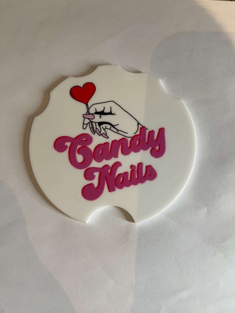 May include: White car coaster with a scalloped edge, featuring a hand holding a red heart-shaped lollipop. The words "Candy Nails" are written in pink and magenta script.