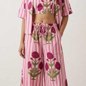 Peut inclure: Ensemble trois pièces à rayures roses et blanches avec un motif floral. L'ensemble comprend une chemise à manches courtes, un crop top et un pantalon large. Le motif floral est magenta foncé avec des feuilles vertes.