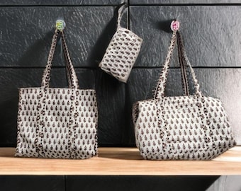 Op de afbeelding: Drie witte en bruine tote tassen met patroon en een kleine etui hangen tegen een donkergrijze muur. De tassen hebben een bladachtig ontwerp en bruine handvatten. De kleine etui hangt aan een haak.