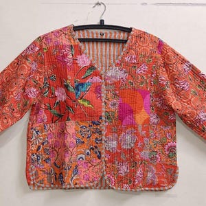 Peut inclure: Veste matelassée à col en V et manches longues. La veste présente un motif patchwork avec des motifs floraux et cachemire dans les tons orange, rose et violet. La veste est suspendue à un cintre noir.