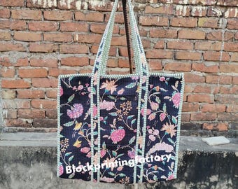Op de afbeelding: Een zwarte tote bag met een bloemenpatroon in roze, geel en groen. De tas heeft een rechthoekige vorm met een gestreepte rand en handvatten. De tekst "blockprintingfactory" staat op de voorkant.