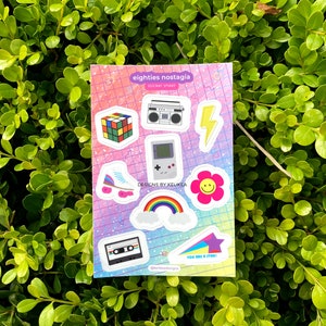 May include: A sticker sheet with a rainbow, a cassette tape, a star, a flower, a roller skate, a game console, a lightning bolt, a boombox, and a Rubik's cube. The sheet has a pink, blue, and purple background with a grid pattern. The text on the sheet reads "eighties nostalgia sticker sheet" and "Designs by Keliikea".