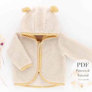 Pode incluir: Uma jaqueta com capuz de cor creme com guarnições quadriculadas amarelas e brancas. A jaqueta tem um fecho de correr e um capuz em forma de orelhas. O texto "PDF Pattern & Tutorial 3 to 12 months" está num círculo branco com um contorno preto.