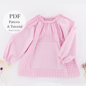 May include: A pink gingham pattern tunic with long sleeves and a front pocket. The text "PDF Pattern & Tutorial From 1 to 4 y/o" is in a white circle. The tunic is designed for children.
