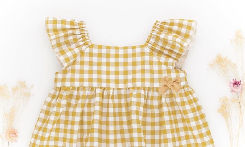K&ouml;nnte beinhalten: Ein gelbes und wei&szlig;es kariertes, &auml;rmelloses Kleid mit einer Schleife auf der Vorderseite. Das Kleid hat einen R&uuml;schenkragen und einen gerafften Rock.