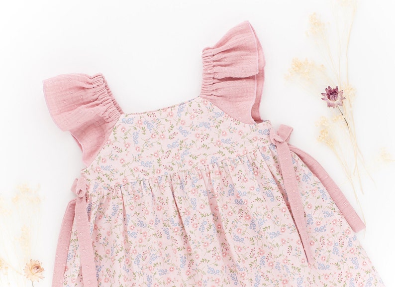 K&ouml;nnte beinhalten: Ein rosa Blumenkleid mit R&uuml;schen&auml;rmeln und Bindeb&auml;ndern. Das Kleid ist aus einem weichen, leichten Stoff gefertigt und hat einen zarten Blumendruck.