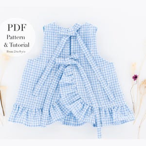 Puede incluir: Top con estampado de cuadros vichy azul claro y blanco con dobladillo con volantes y detalle de lazo. El top es un patrón PDF y un tutorial para niños de 2 a 8 años. Flores secas dispuestas a ambos lados.