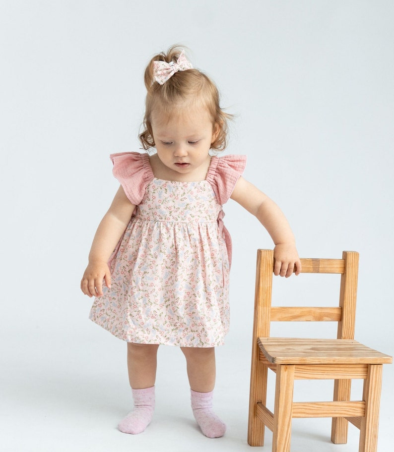 K&ouml;nnte beinhalten: Ein kleines Kind tr&auml;gt ein rosa Blumenkleid mit R&uuml;schen&auml;rmeln und steht vor einem Holzstuhl. Das Kind tr&auml;gt rosa Socken und eine rosa Schleife im Haar.