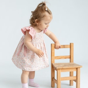 K&ouml;nnte beinhalten: Ein kleines Kind in einem rosa Blumenkleid mit R&uuml;schen&auml;rmeln steht neben einem kleinen Holzstuhl. Das Kind schaut auf den Stuhl.