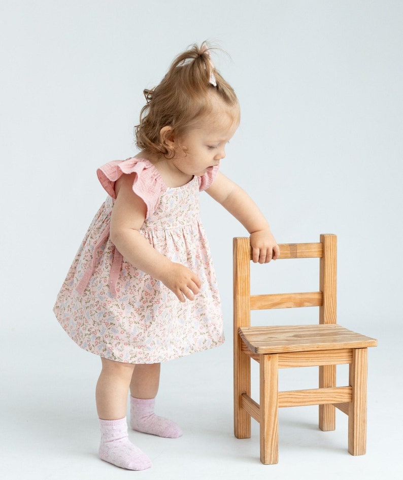 K&ouml;nnte beinhalten: Ein kleines Kind in einem rosa Blumenkleid mit R&uuml;schen&auml;rmeln steht neben einem kleinen Holzstuhl. Das Kind schaut auf den Stuhl.