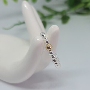 Puede incluir: Un anillo de cuentas de plata con una sola cuenta de oro. El anillo está en un expositor blanco con forma de mano.