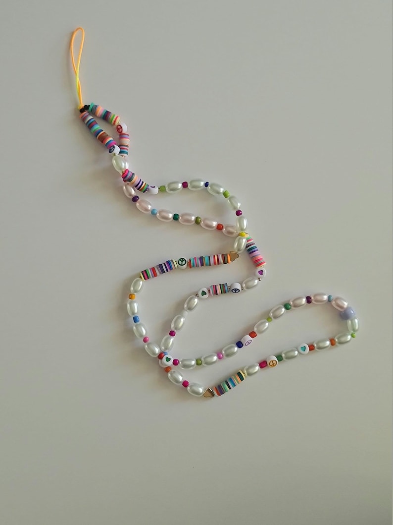 Puede incluir: Una correa de tel&eacute;fono con cuentas de colores con cuentas de perlas blancas, dijes de signo de paz y un dije de coraz&oacute;n. La correa est&aacute; hecha de cuentas rayadas de colores y tiene un lazo de cuerda amarillo en la parte superior.