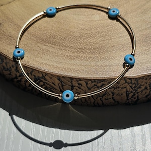 May include: Gold-toned bangle bracelet featuring six light blue and black evil eye beads. The bracelet is made of two parallel curved metal bars. The beads are evenly spaced around the bracelet.