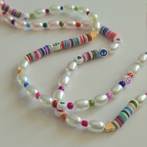 Puede incluir: Un collar de varias hebras con cuentas de perlas blancas y cuentas de colores con signos de paz y corazones. El collar est&aacute; en capas y tiene un estilo bohemio.