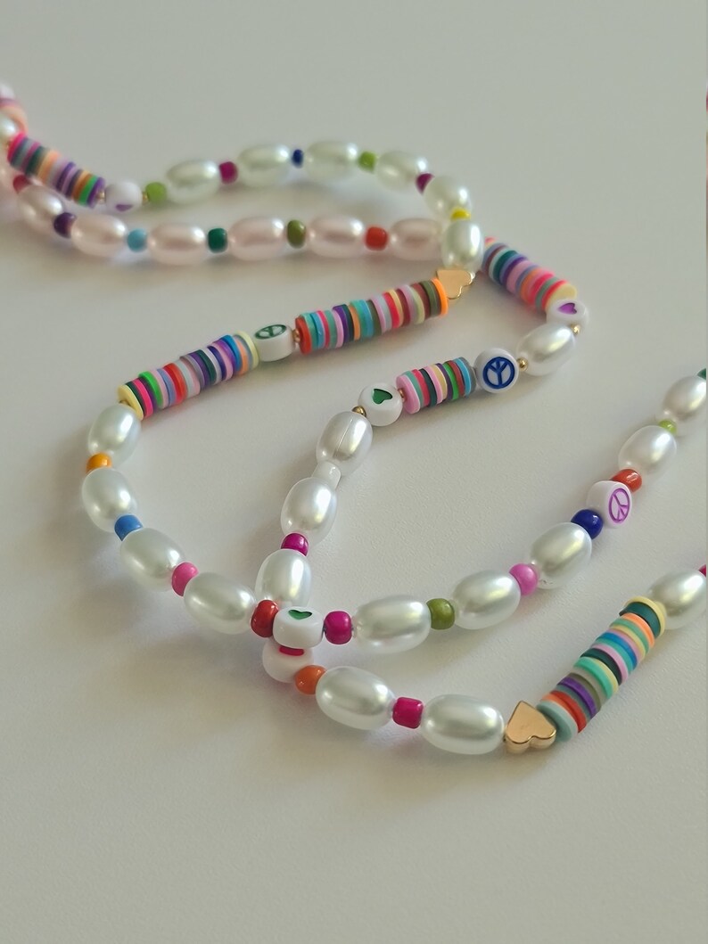 Puede incluir: Un collar de varias hebras con cuentas de perlas blancas y cuentas de colores con signos de paz y corazones. El collar est&aacute; en capas y tiene un estilo bohemio.