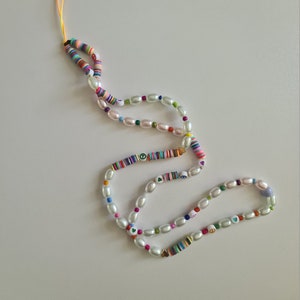Puede incluir: Un abalorio para tel&eacute;fono colorido con cuentas de perlas blancas, dijes de signo de paz y coraz&oacute;n, y un patr&oacute;n de rayas arco&iacute;ris.