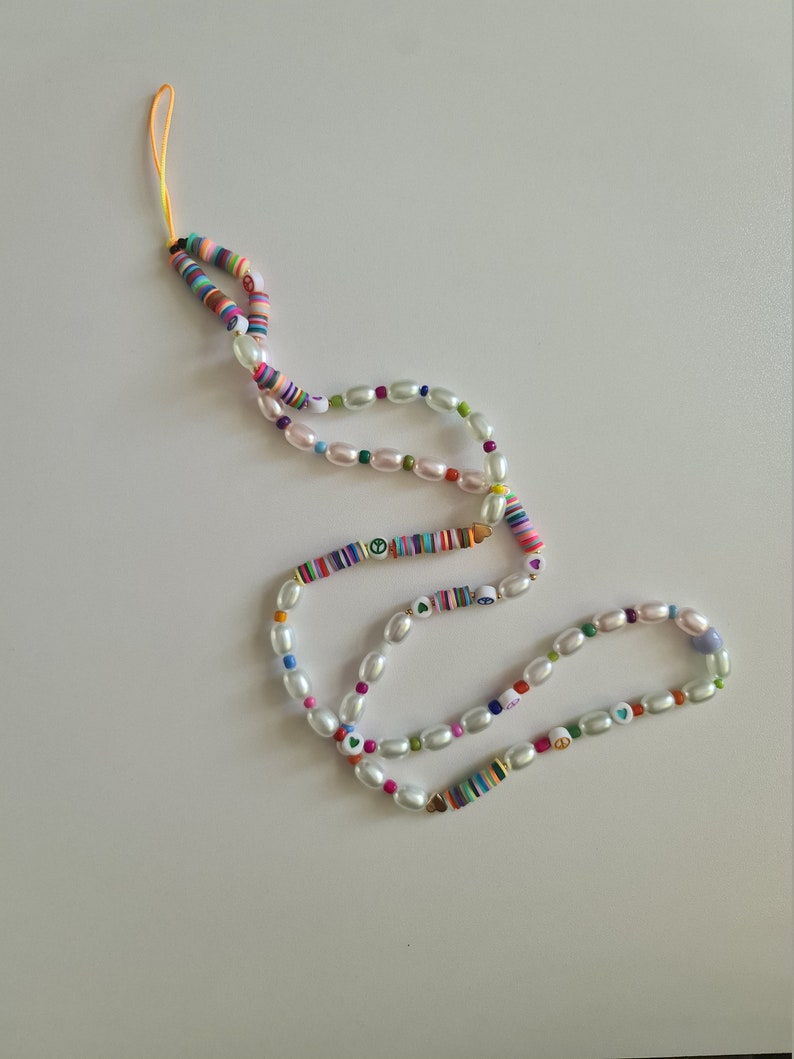 Puede incluir: Un abalorio para tel&eacute;fono colorido con cuentas de perlas blancas, dijes de signo de paz y coraz&oacute;n, y un patr&oacute;n de rayas arco&iacute;ris.
