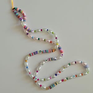 Puede incluir: Un abalorio para tel&eacute;fono colorido con perlas blancas, signos de paz y corazones. El abalorio est&aacute; hecho de cuentas de colores y tiene una cuerda amarilla.