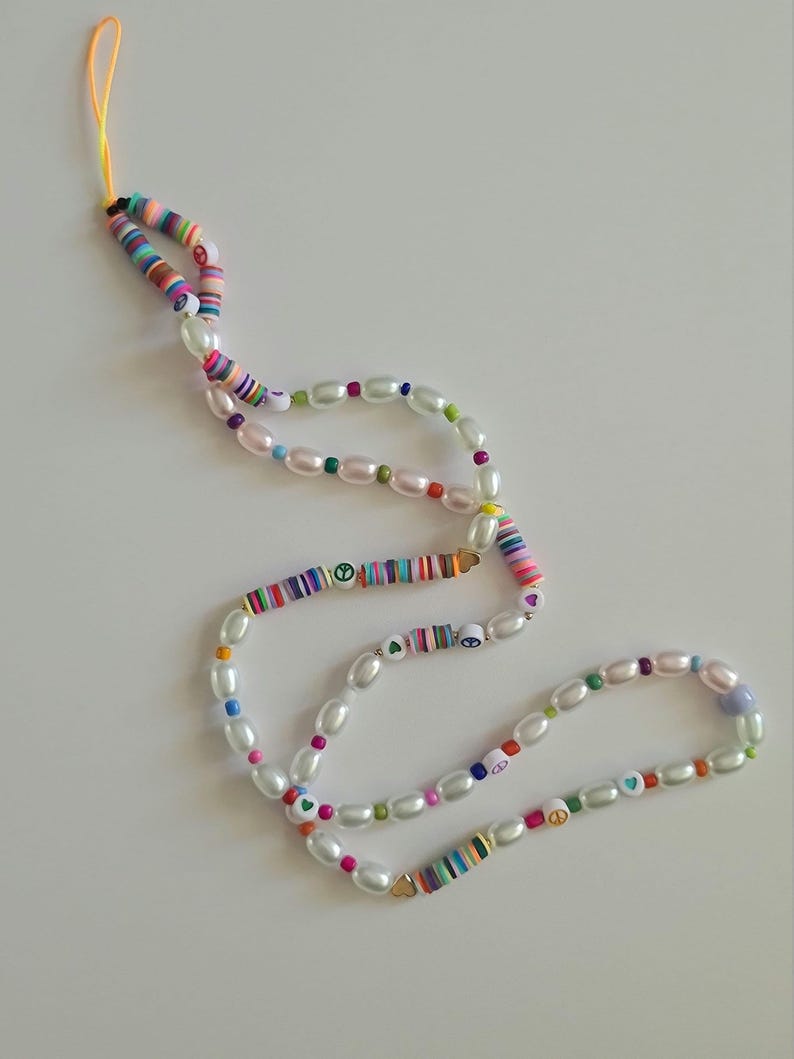 Puede incluir: Un abalorio para tel&eacute;fono colorido con perlas blancas, signos de paz y corazones. El abalorio est&aacute; hecho de cuentas de colores y tiene una cuerda amarilla.