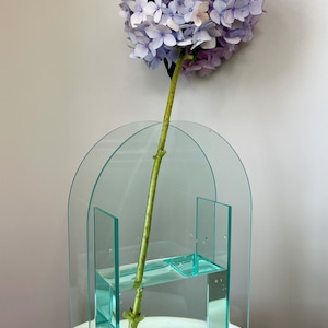 May include: A clear glass vase with an arched top and a rectangular base. The vase is filled with water and a single stem of light purple hydrangeas.