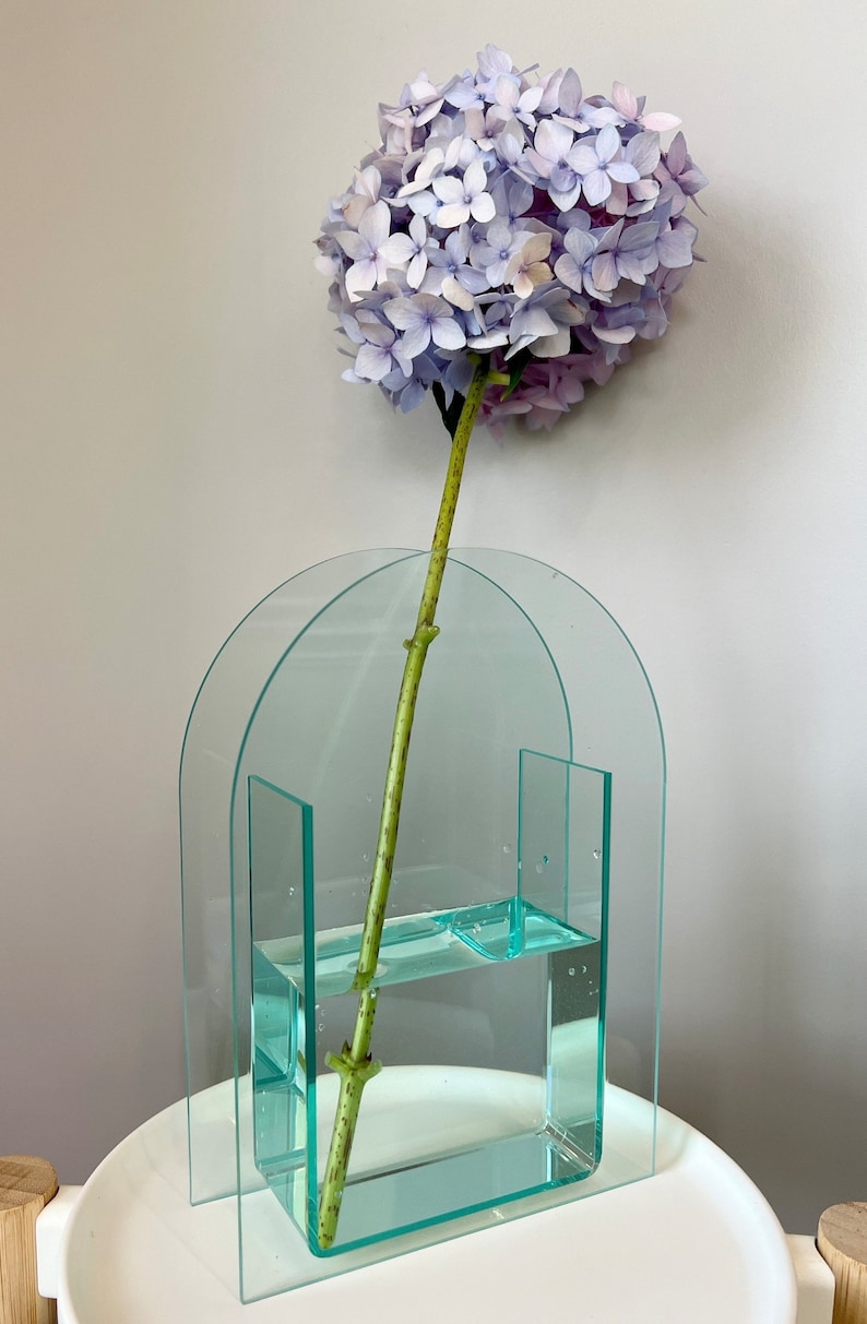 May include: A clear glass vase with an arched top and a rectangular base. The vase is filled with water and a single stem of light purple hydrangeas.