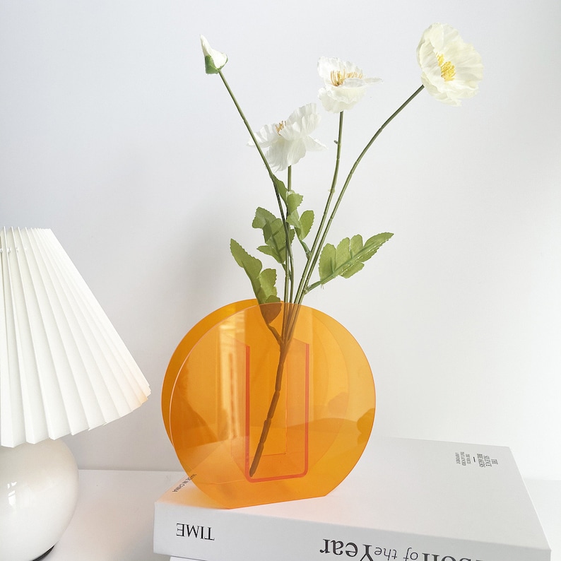 May include: A round, orange acrylic vase with a clear rectangular opening in the center. The vase is filled with white flowers and green stems.