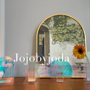 Puede incluir: Cinco jarrones de vidrio iridiscente con diseños geométricos. Los jarrones están dispuestos sobre una mesa de madera frente a una imagen enmarcada con un marco dorado. La imagen muestra un paisaje con una pared blanca y una vid verde.