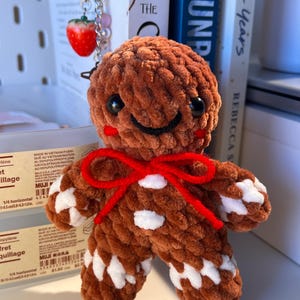 May include: A handmade brown crocheted gingerbread man with white icing details and a red bow. The plush toy has black button eyes and a smiling face. A strawberry charm hangs in the background. The gingerbread man is a festive holiday decoration.